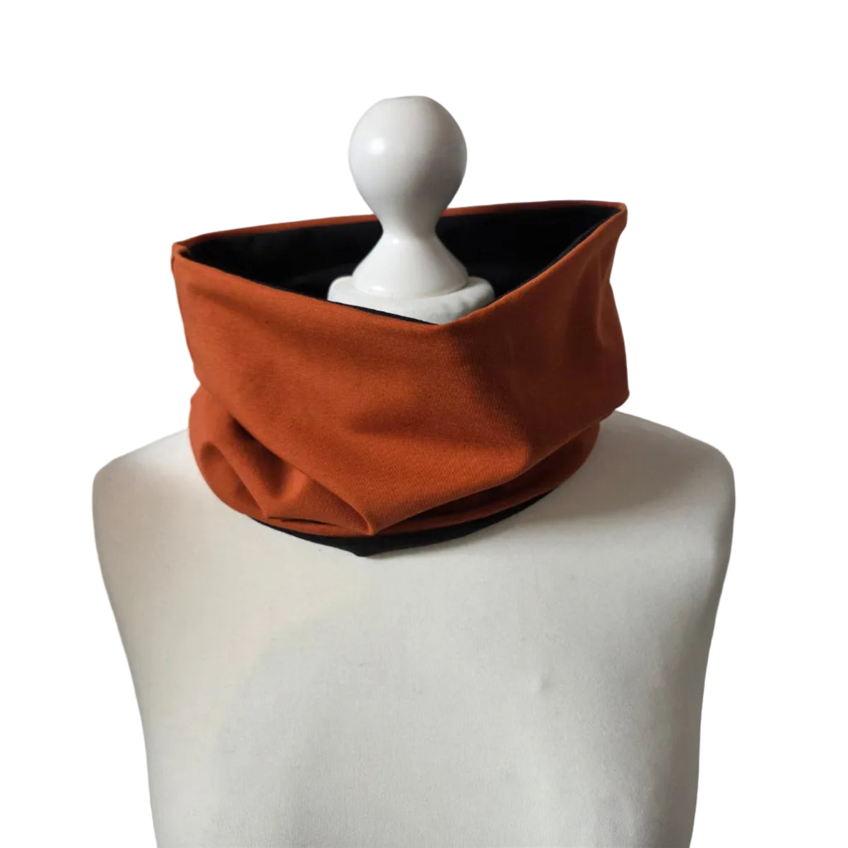An orange-brown fabric neck warmer or cowl with a black inner lining, displayed on a white mannequin torso against a white brick wall background.