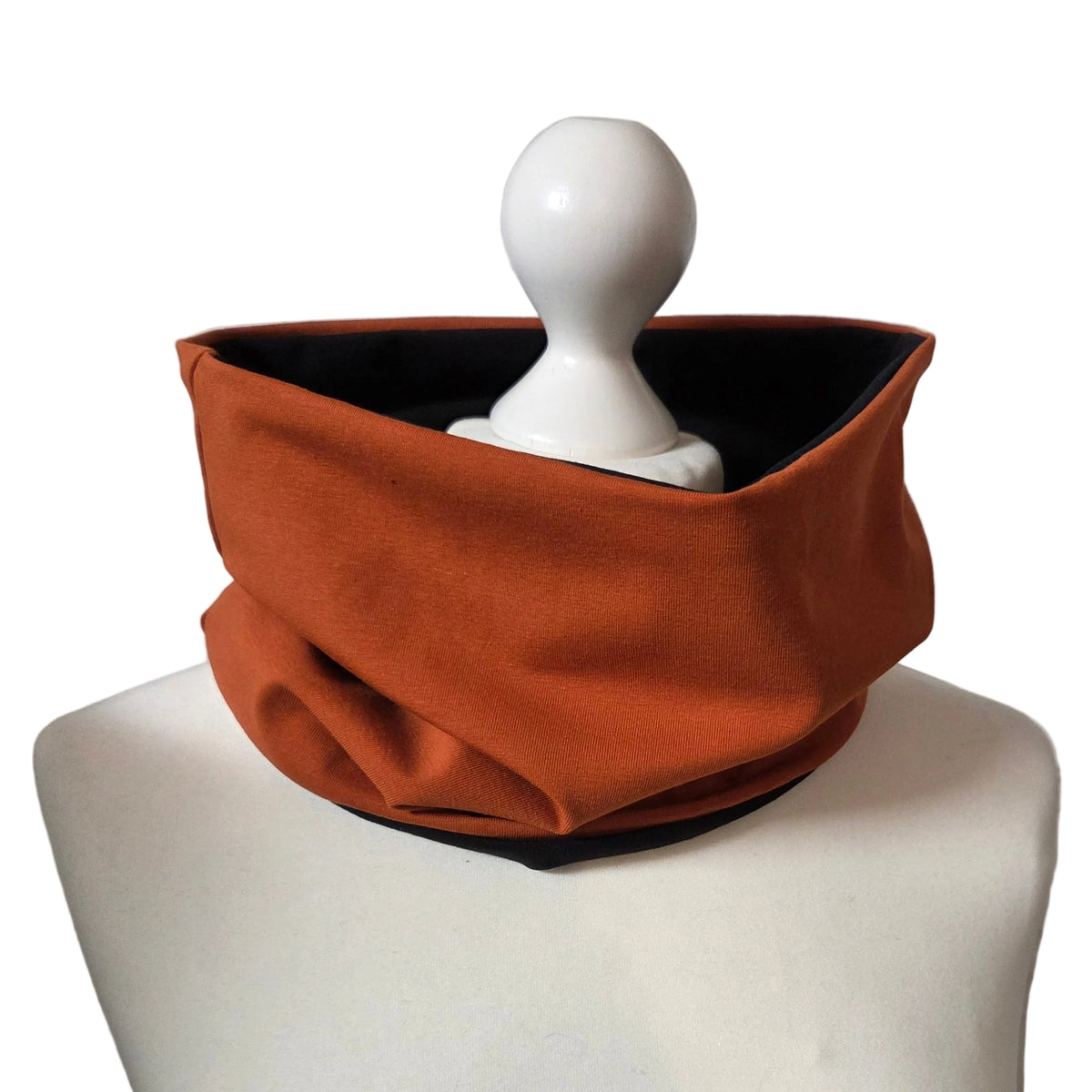 An orange-brown fabric neck warmer or cowl displayed on a white mannequin torso against a white brick wall background.