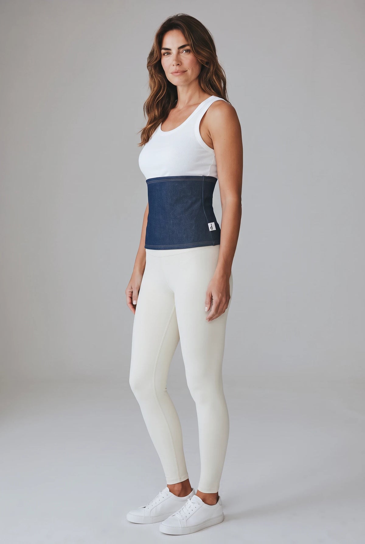 A woman wearing a Fly and Fit dark blue denim-look fleece core warmer over a white tank top and cream leggings.